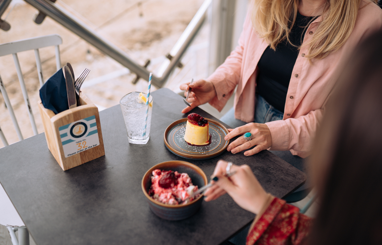 Two women sat at a table in the outside seating area of a restaurant with desserts and beach views