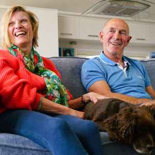 A couple sat smiling together on a sofa with a dog