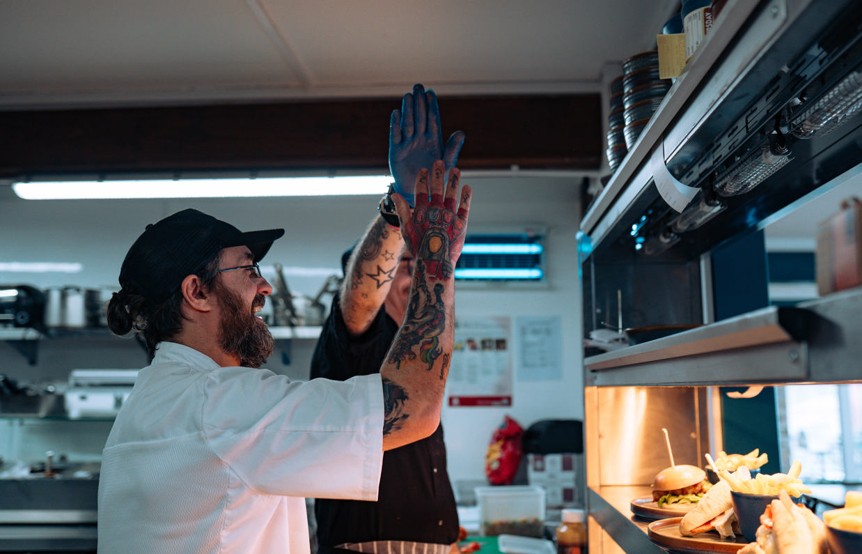 Two male chefs with tattoos in the kitchen of Drift Bar & Grill high fiving behind the serving counter after working together to plate up dishes
