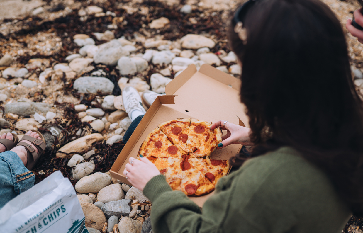 A woman sat on the beach with a pizza box open on her lap picking up a slice