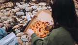A woman sat on the beach with a pizza box open on her lap picking up a slice