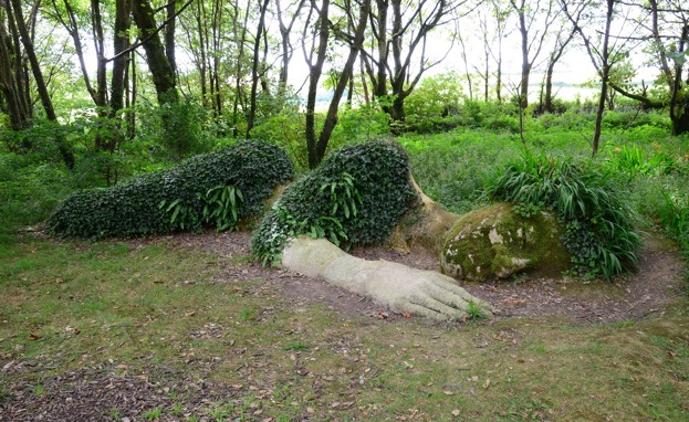 A bigger than life size stone sculpture of a human laying down with greenery growing over it to mimic clothing and hair