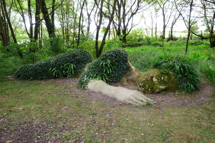 A bigger than life size stone sculpture of a human laying down with greenery growing over it to mimic clothing and hair