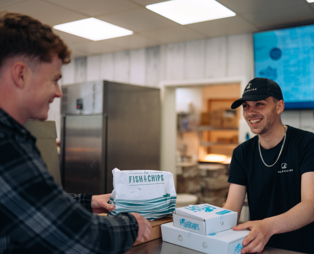 Two young men in a fish and chip takeaway both smiling whilst the server passes the customer takeaway boxes