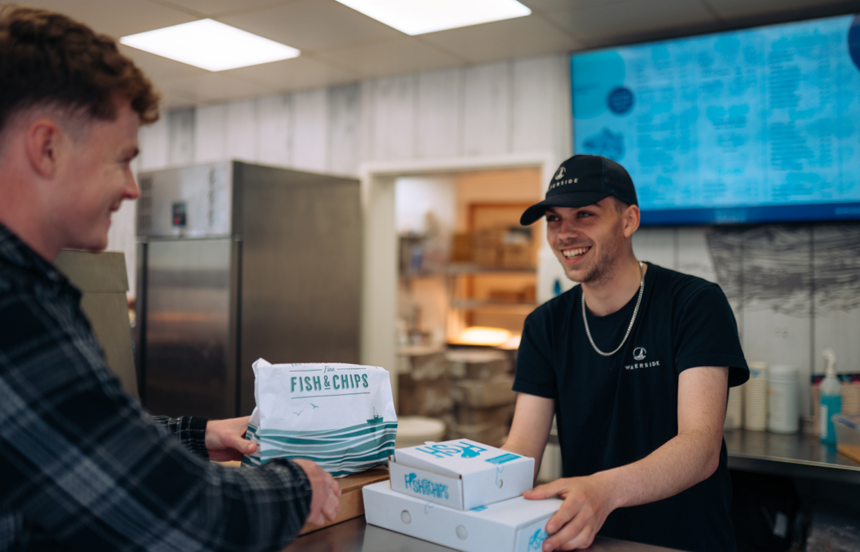 Two young men in a fish and chip takeaway both smiling whilst the server passes the customer takeaway boxes