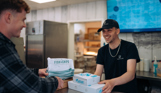 Two young men in a fish and chip takeaway both smiling whilst the server passes the customer takeaway boxes
