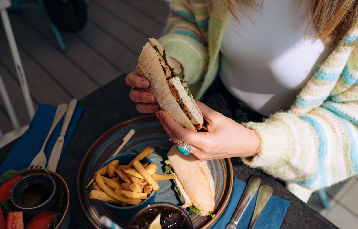 A filled baguette being held by a lady above a plate of chips 