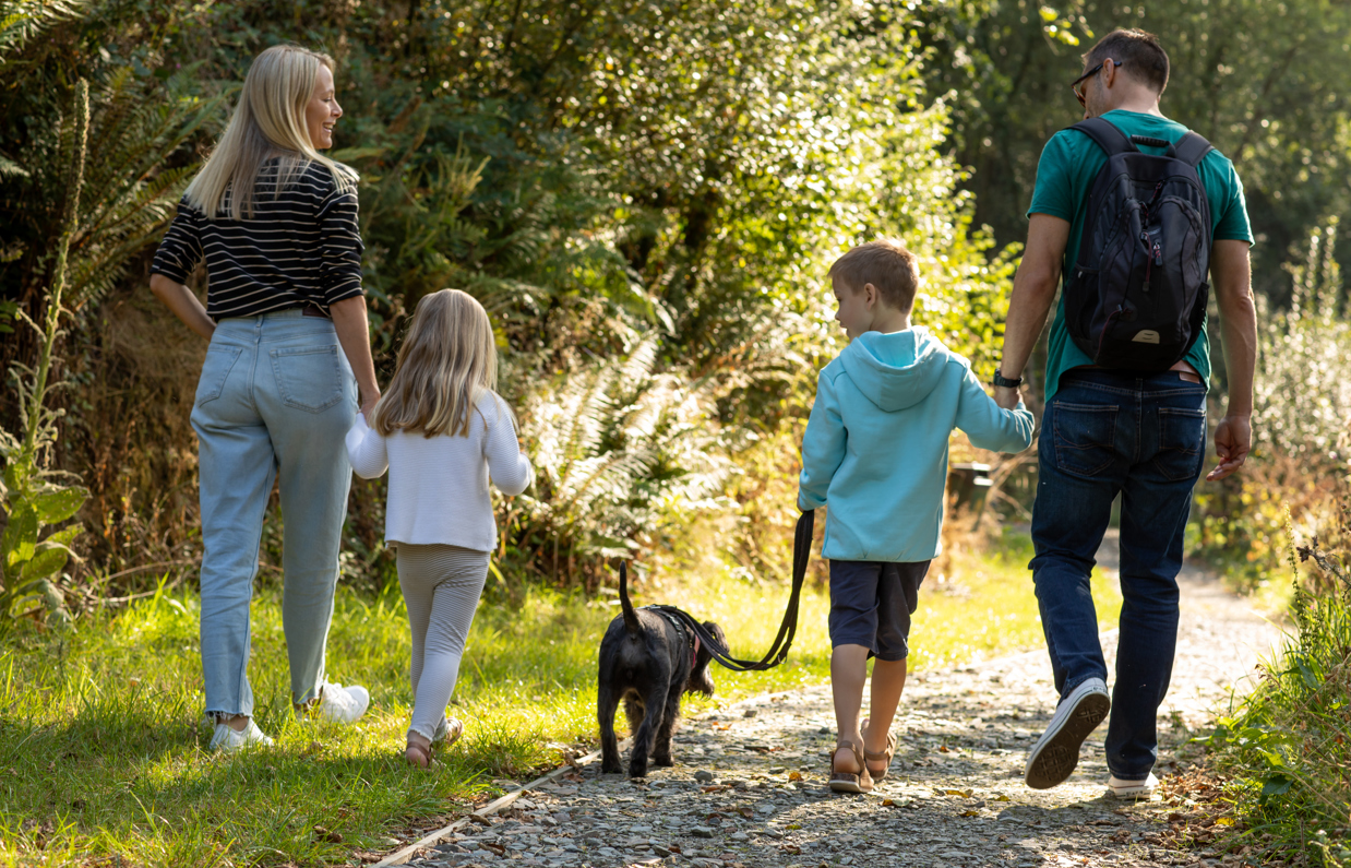 A family of four walking through the woods on a nature trail with a small black dog on a sunny day