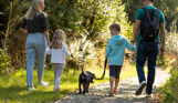 A family of four walking through the woods on a nature trail with a small black dog on a sunny day