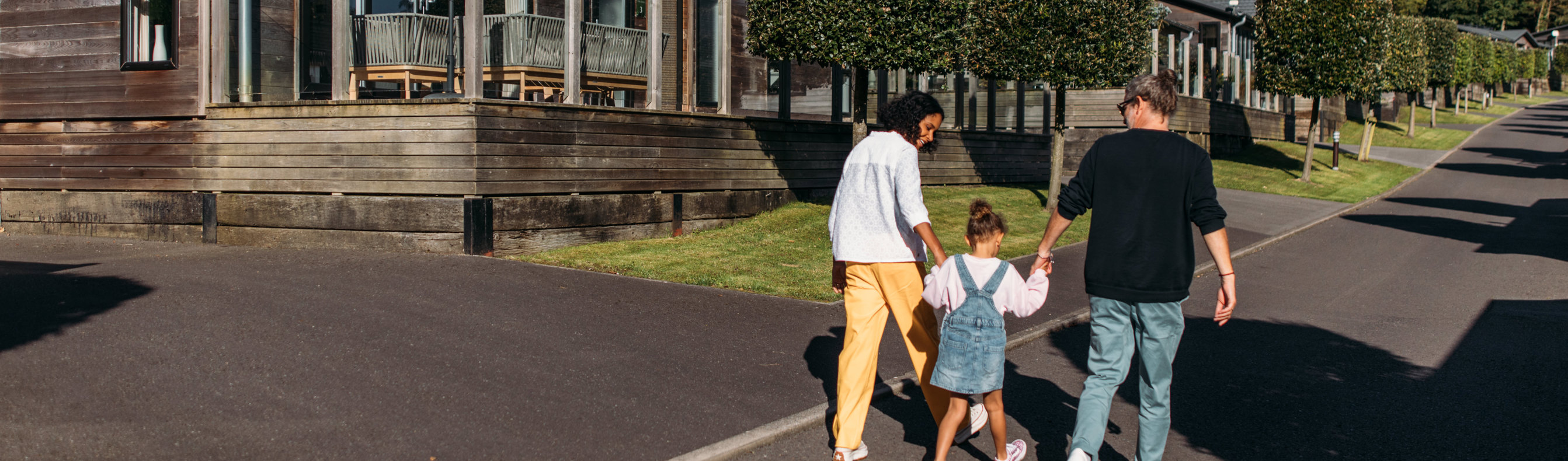 A family of three walking down a road on a lodge park surrounded by holiday lodges and woodland on a sunny, blue sky day