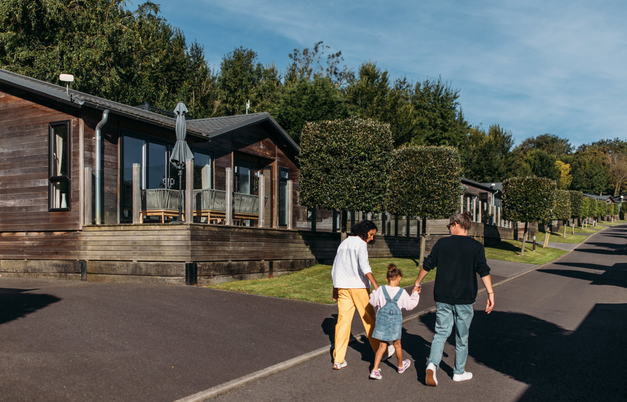 A family of three walking down a road on a lodge park surrounded by holiday lodges and woodland on a sunny, blue sky day