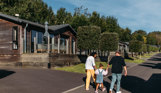A family of three walking down a road on a lodge park surrounded by holiday lodges and woodland on a sunny, blue sky day