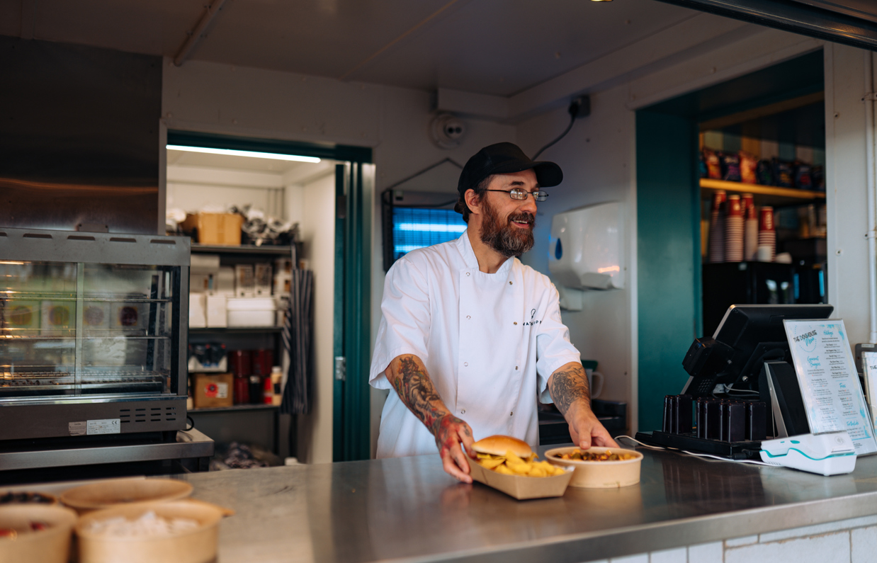 A male server at a street food shack smiling serving some food across the counter
