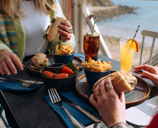Two women sat at a table in the outside seating area of a restaurant with filled baguettes, chips, salad, drinks and seaviews