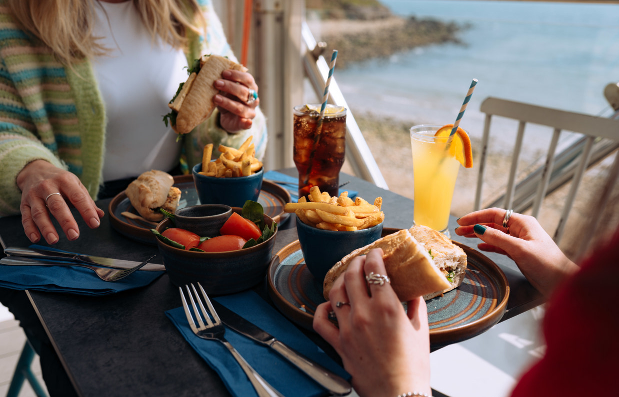 Two women sat at a table in the outside seating area of a restaurant with filled baguettes, chips, salad, drinks and seaviews