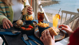 Two women sat at a table in the outside seating area of a restaurant with filled baguettes, chips, salad, drinks and seaviews