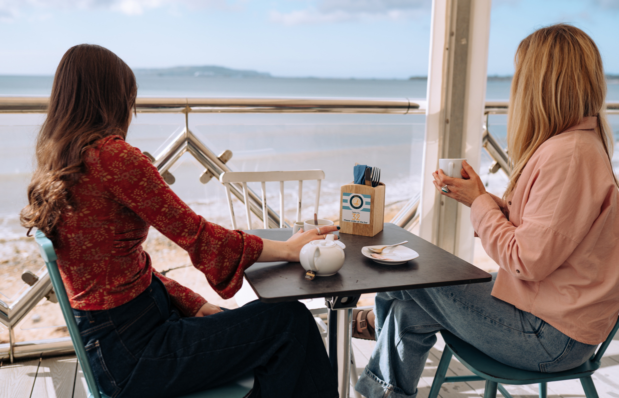 Two women sat at a table at the outside seating covered area of Drift Bar & Grill looking out towards the sea views with a hot drink