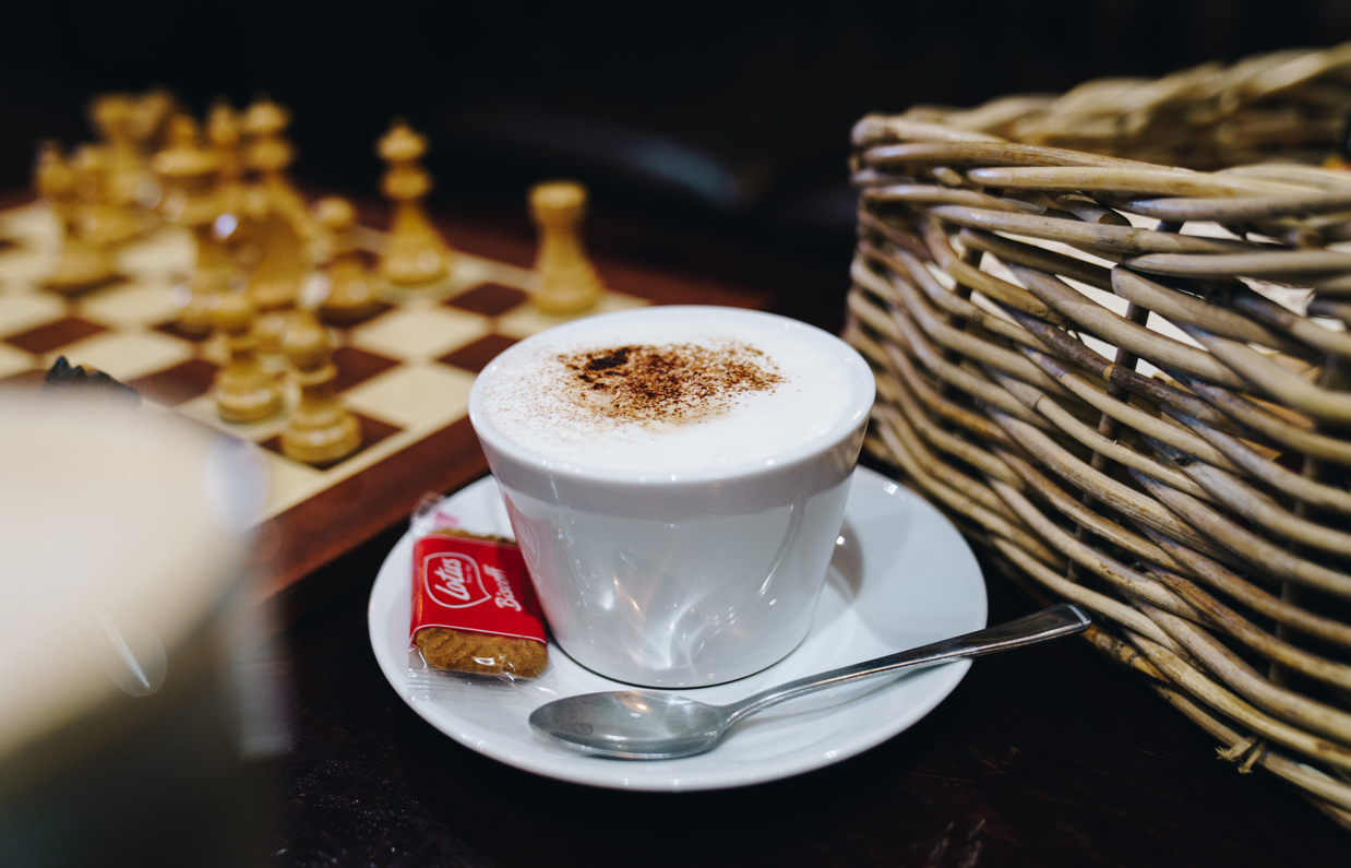 A milky coffee in a teacup and saucer with a biscuit and a chess board