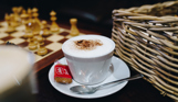 A milky coffee in a teacup and saucer with a biscuit and a chess board