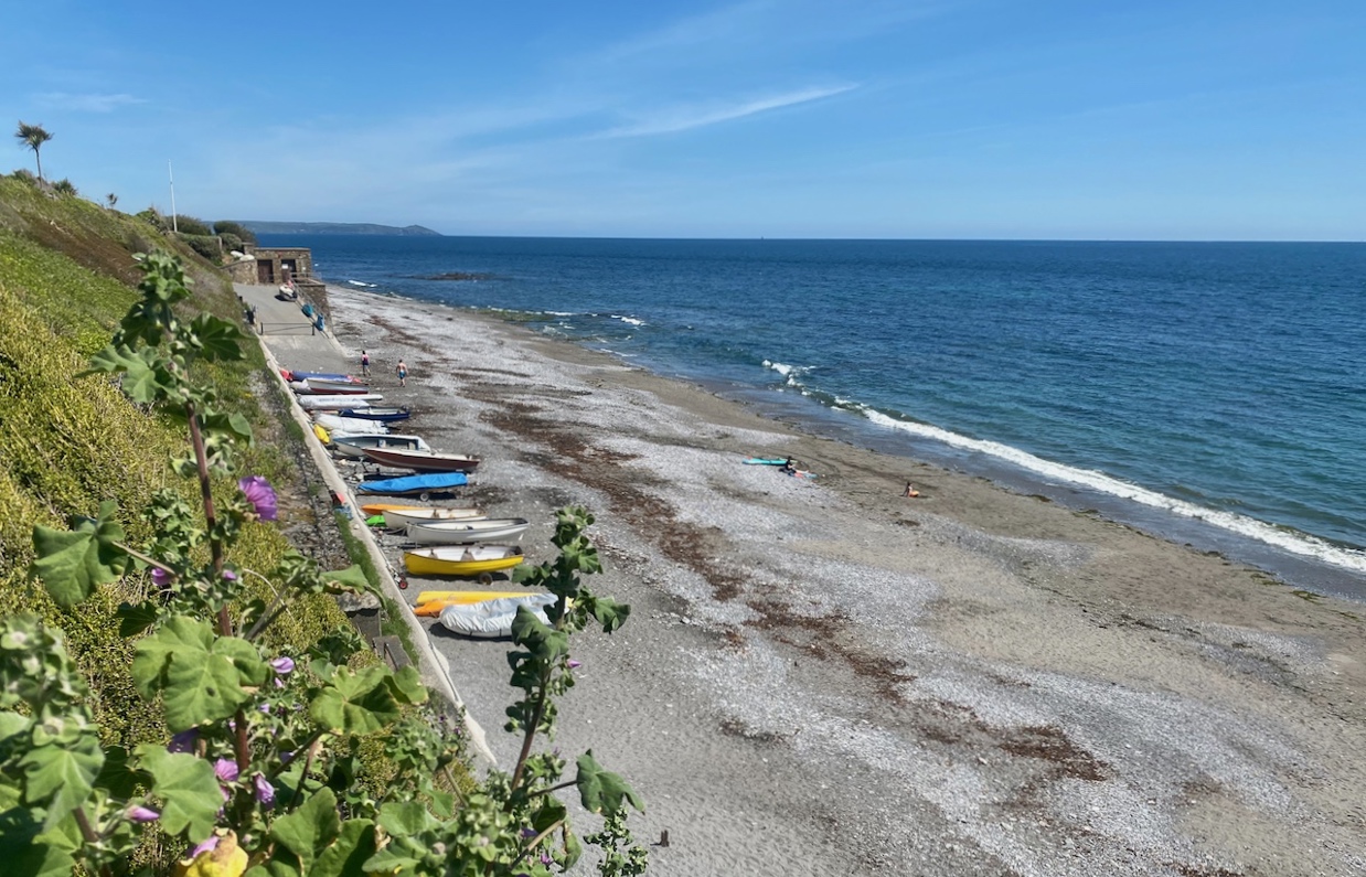 A small shingle beach at the bottom of rolling countryside hills on a sunny, blue sky day with various small boats on the shore