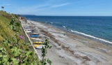 A small shingle beach at the bottom of rolling countryside hills on a sunny, blue sky day with various small boats on the shore