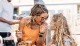 A mother and daughter smiling gleefully at each other at an outside table with street food and drinks with a chef behind them serving food