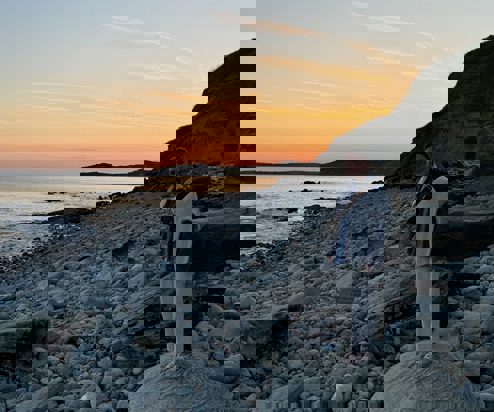 A lady standing on a large rock on a stoney beach at sunset