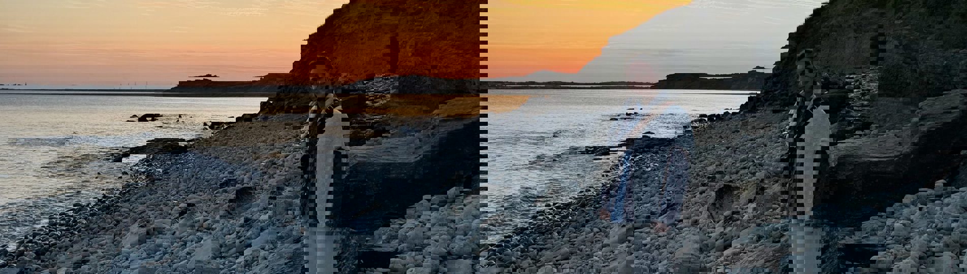 A lady standing on a large rock on a stoney beach at sunset