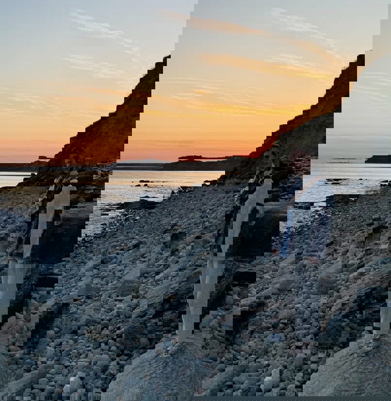 A lady standing on a large rock on a stoney beach at sunset