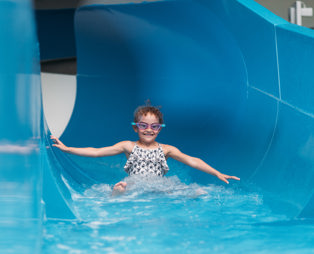 A young girl wearing goggles going down a blue indoor waterslide with a grin on her face