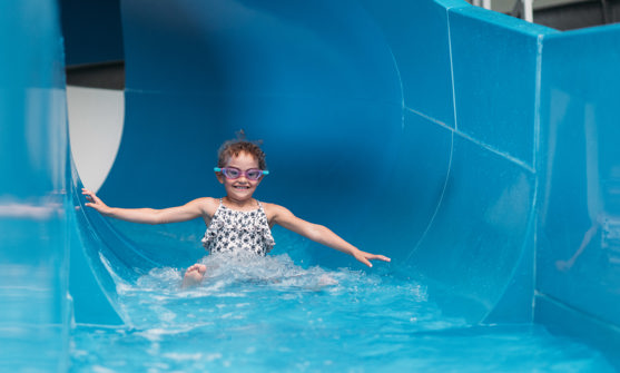 A young girl wearing goggles going down a blue indoor waterslide with a grin on her face