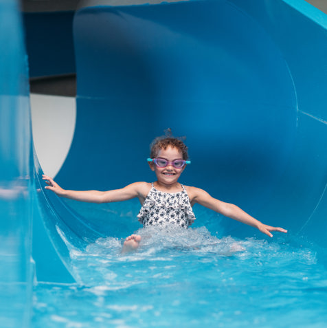A young girl wearing goggles going down a blue indoor waterslide with a grin on her face