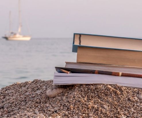 A stack of books on shingle beach with a ship in the sea in the background