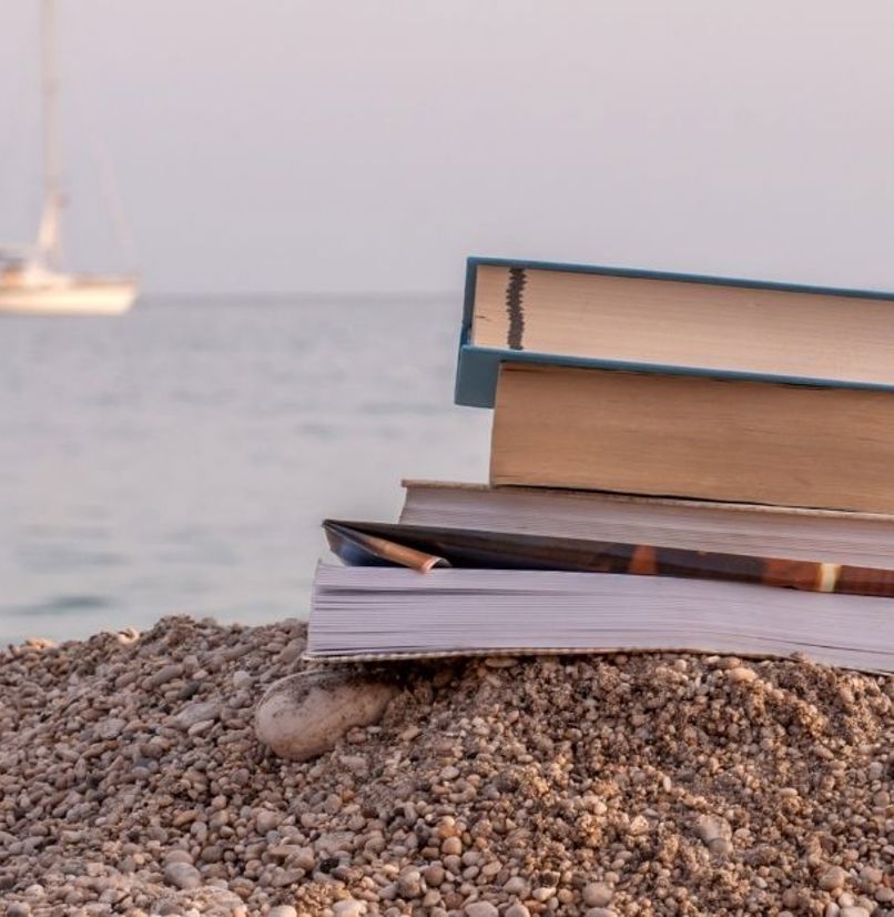 A stack of books on shingle beach with a ship in the sea in the background