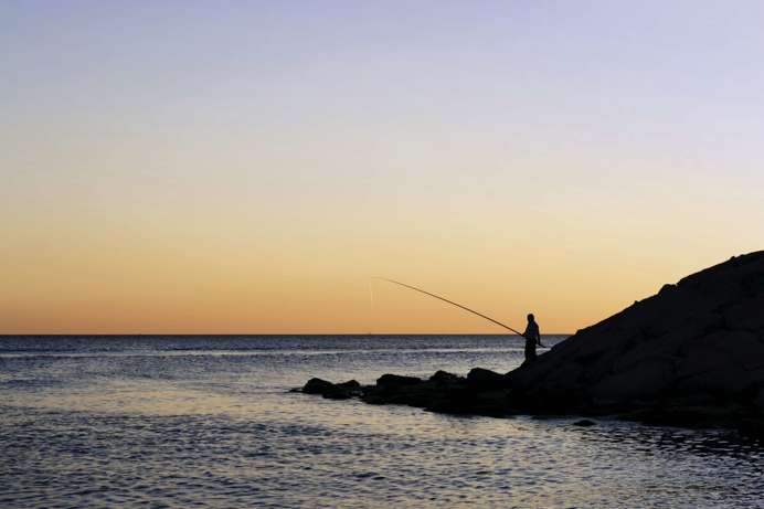 A silhouette of a person fishing in the sea with tranquil water and striking sky