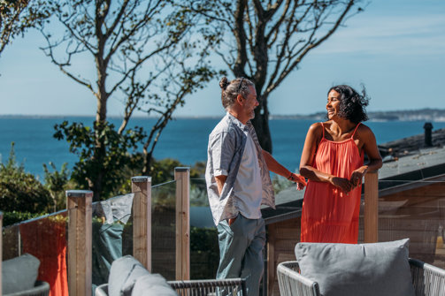Coupe laughing together on the deck of a lodge with view of coast behind them