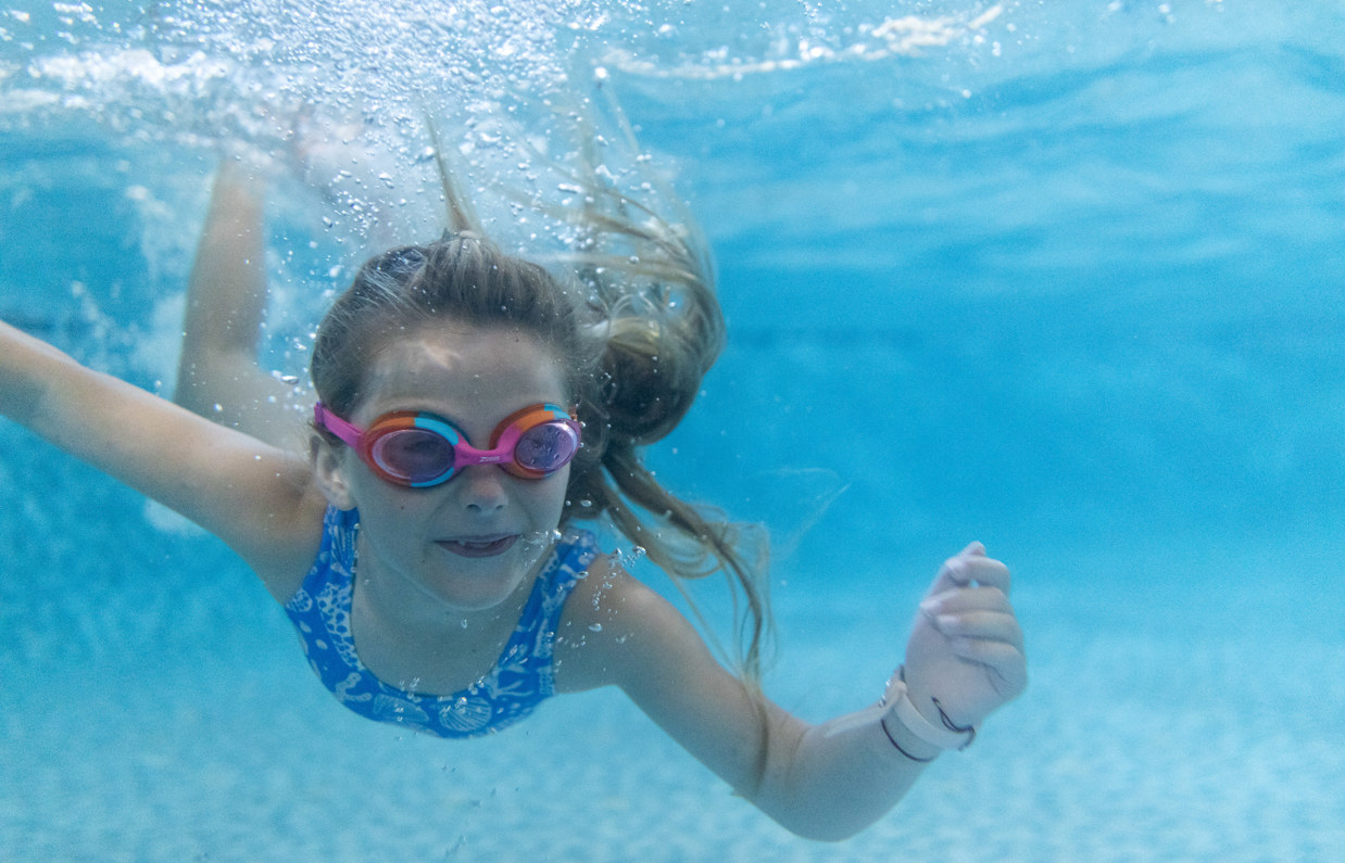 A young girl wearing goggles swimming underwater in a heated outdoor swimming pool