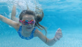 A young girl wearing goggles swimming underwater in a heated outdoor swimming pool