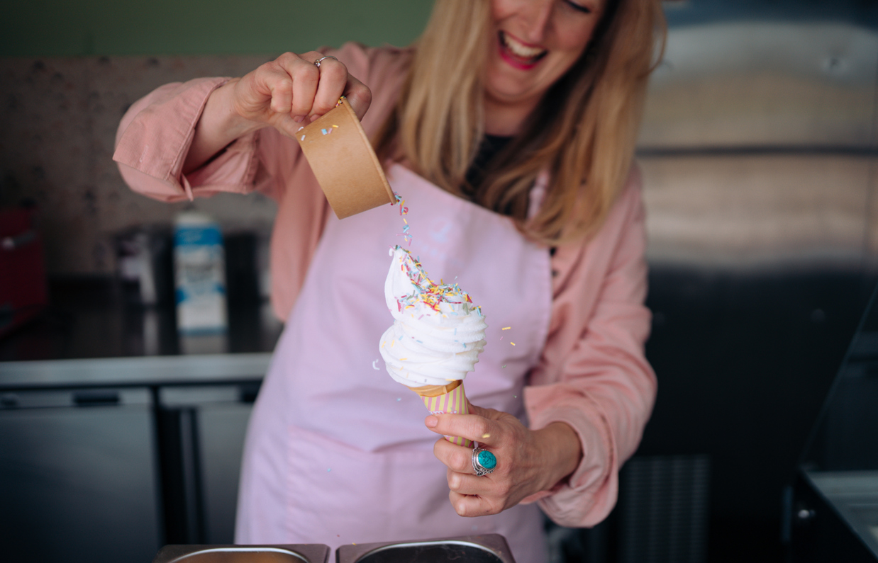 A Mr Whippy style ice cream having sprinkles poured over the top with some sprinkles flying off whilst the woman holding it smiles happily
