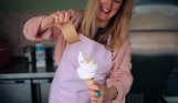 A Mr Whippy style ice cream having sprinkles poured over the top with some sprinkles flying off whilst the woman holding it smiles happily