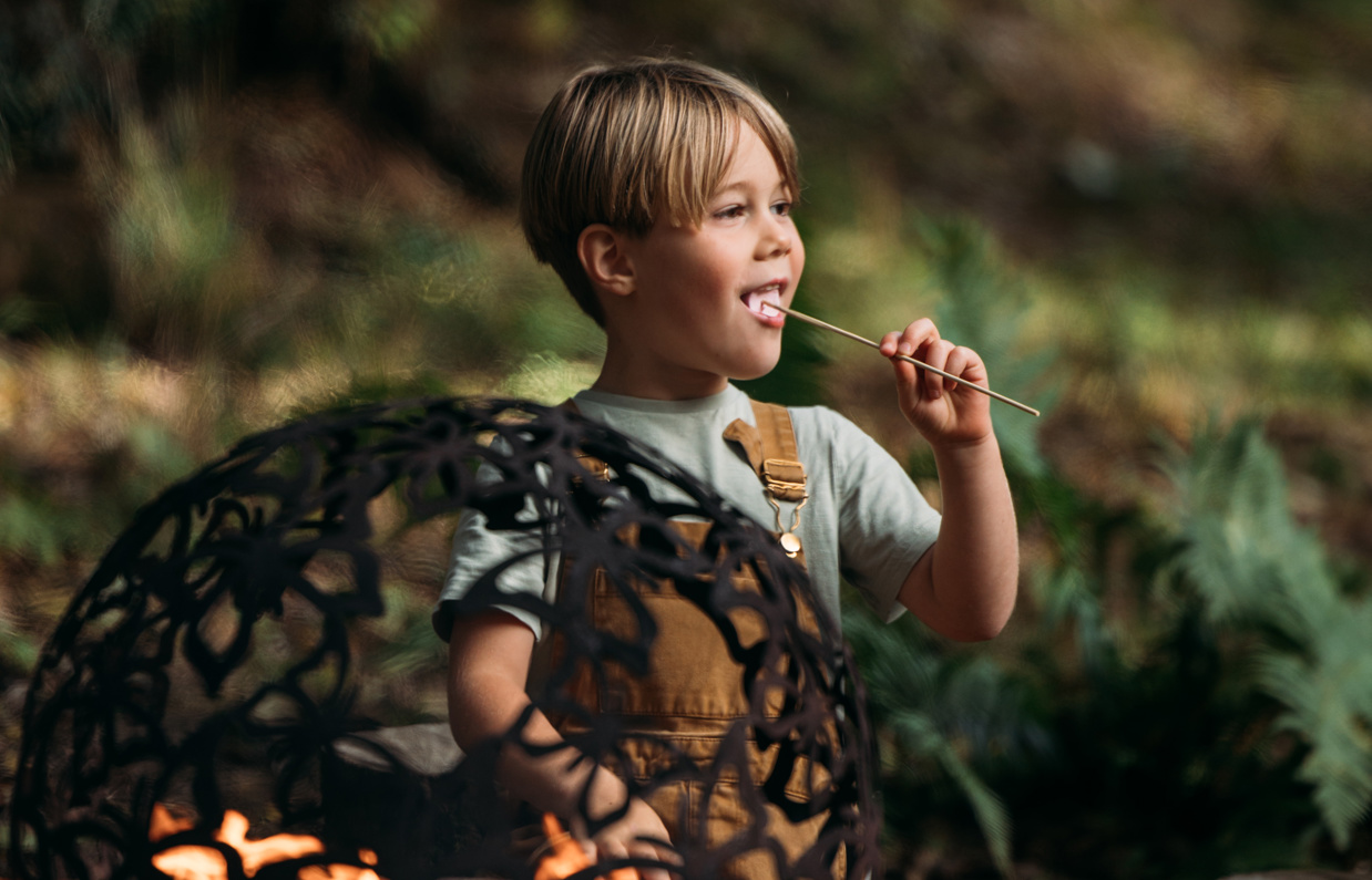 A young boy kneeling down in the woods beside a campfire with a toasted marshmallow in his mouth