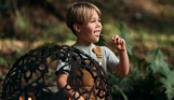 A young boy kneeling down in the woods beside a campfire with a toasted marshmallow in his mouth