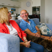 A couple sat smiling together on a sofa with a dog
