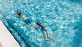 A young boy underwater in an outdoor pool on an aqua jet with a young girl holding onto his leg behind him
