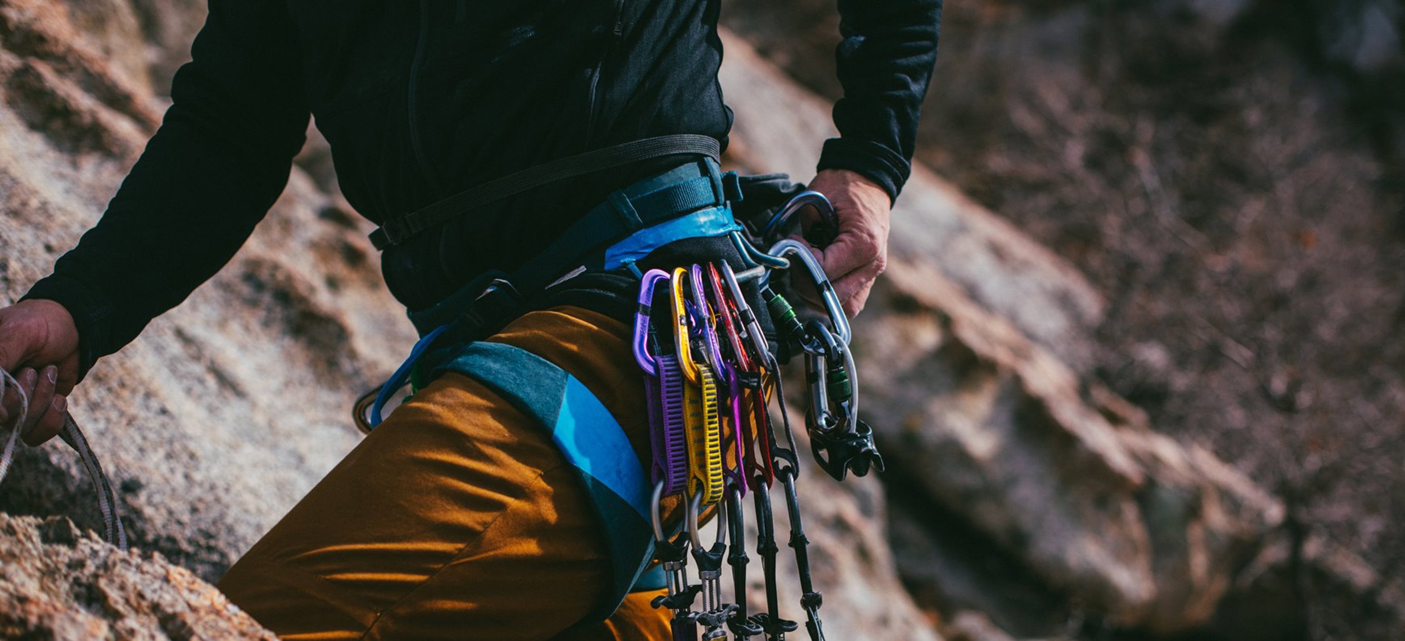 A man's body against a cliff face rock climbing with various ropes, equipment and harnesses round his waist