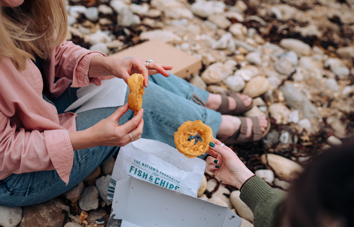Two women sat on a beach with fish and chip takeaway picking up onion rings