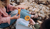 Two women sat on a beach with fish and chip takeaway picking up onion rings