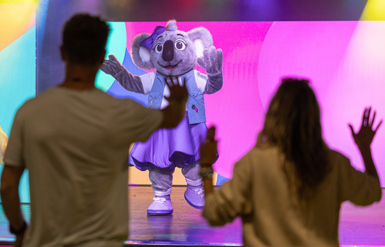 A koala mascot, Kimi on stage in an entertainment venue with a couple copying her dancing