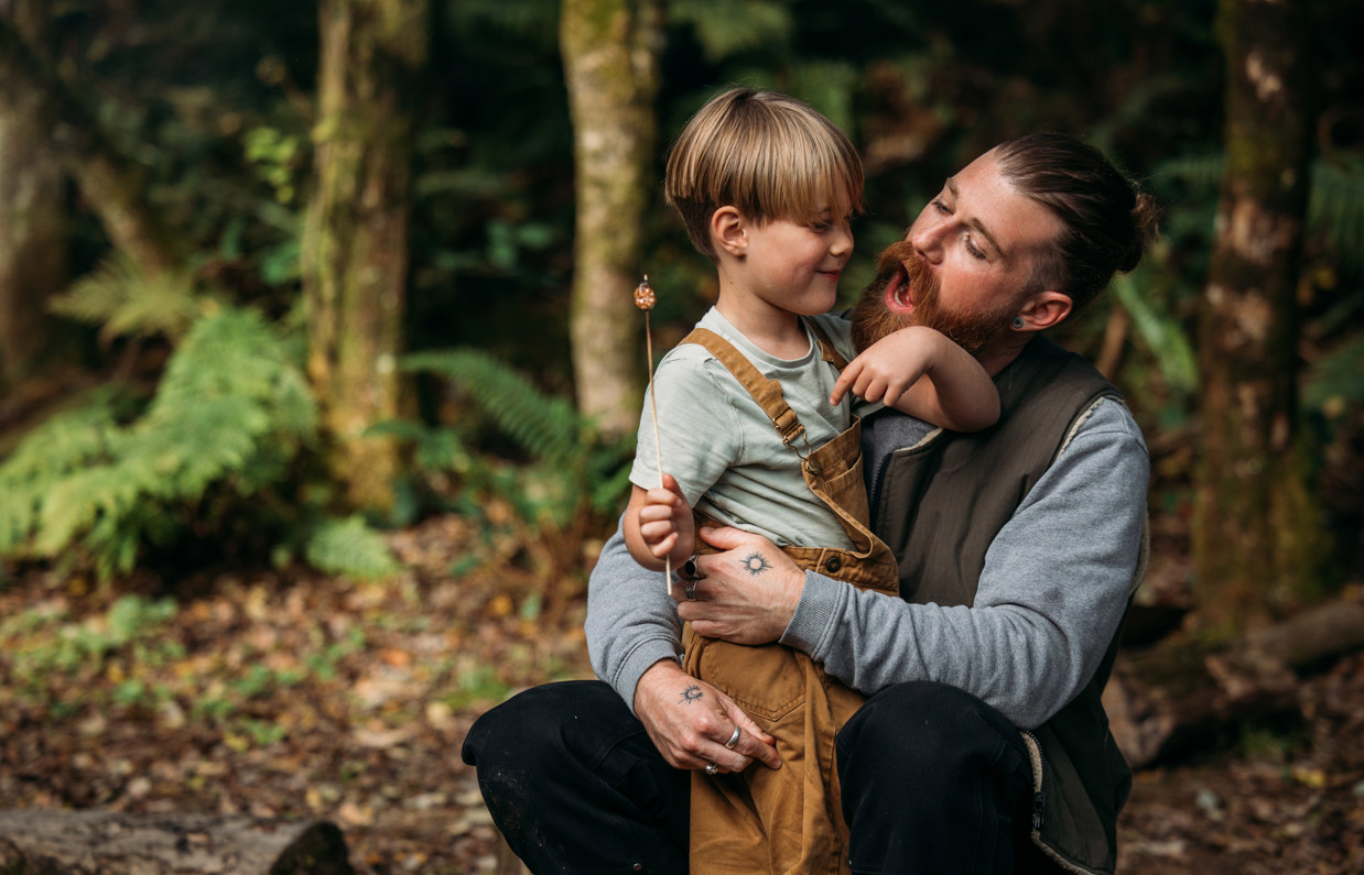 A young boy and his dad smiling together in the woods with toasted marshmallows