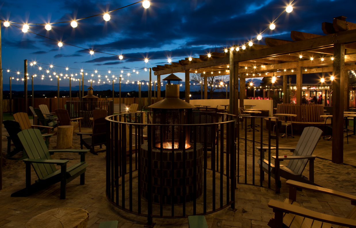 An outdoor seating area with a fire pit and chairs and fairy lights under a veranda at night